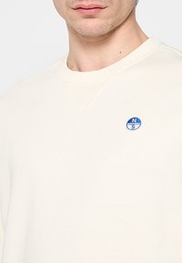 Felpa color crema con scollo rotondo, caratterizzata da un piccolo logo rotondo blu sul lato sinistro del petto e dettagli di cucitura visibili.