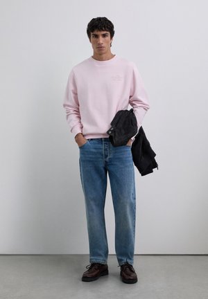 Jonge man staat in een minimalistische studio, gekleed in een lichtroze sweatshirt, blauwe jeans en donkerbruine schoenen, en houdt een zwarte jas vast.