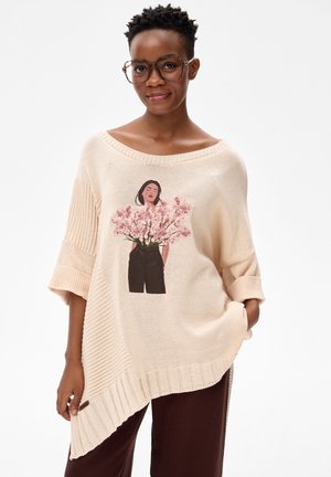Kobieta ubrana w beżowy asymetryczny sweter z dzianiny z ilustracją kobiety trzymającej różowe kwiaty na przodzie, ciemne spodnie i okulary.