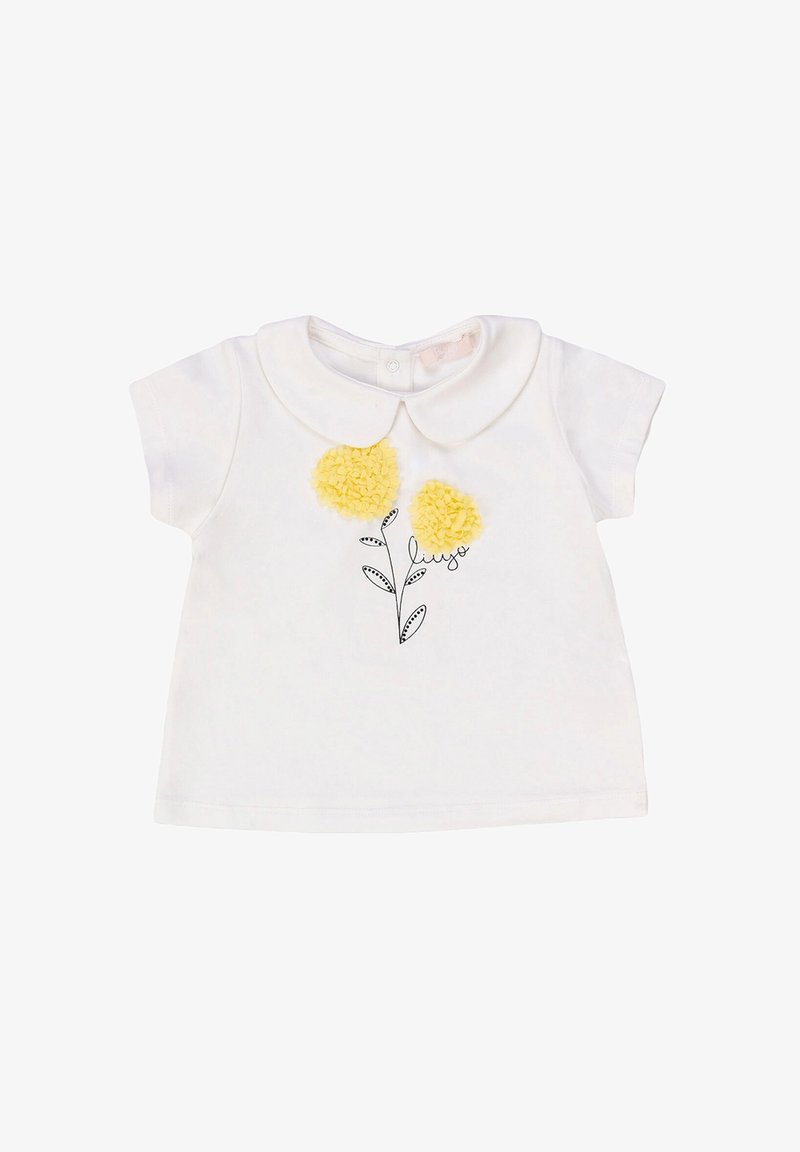 Camicia bianca in cotone a maniche corte con colletto Peter Pan, caratterizzata da tre fiori a pom-pom gialli e un'illustrazione di foglia nera sottostante.