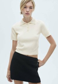 Strikket beige polo-top med korte ærmer og en struktureret krave, dækket med en sort mini-nederdel med en sidelomme og knapdetalje.