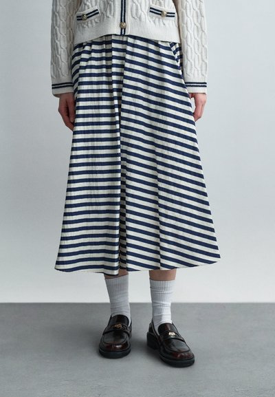 Vue de la taille aux pieds d'une personne portant une jupe midi rayée bleu marine et blanche, un cardigan en tricot écru, des chaussettes blanches et des mocassins noirs sur un sol gris.
