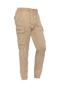 Beige cargobroek met een elastische tailleband en een trekkoord. Beschikt over meerdere zakken, taps toelopende pijpen en een zachte katoenmix stof.