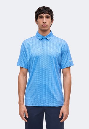 Mężczyzna stoi przodem, ubrany w niebieską wzorzystą koszulkę polo Nike z krótkim rękawem i ciemne spodnie, neutralny wyraz twarzy, jednolite jasne tło.
