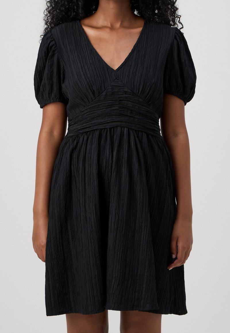 Femme portant une robe noire texturée longueur genou avec des manches bouffantes, un décolleté en V et une taille cintrée, sur un fond uni.