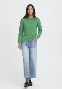 Mujer sonriendo, vistiendo un suéter de punto verde, jeans de un azul claro, calcetines grises y mocasines negros, de pie contra un fondo blanco liso.