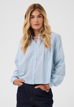 Femme blonde aux cheveux ondulés portant une chemise bleu clair boutonnée et un jean foncé, les mains dans les poches, sur un fond uni.