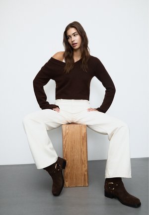 Maglione a maglia marrone con spalle scoperte, jeans a gamba larga avorio e stivali alla caviglia in suede marrone con dettagli in oro, seduta su un blocco di legno.