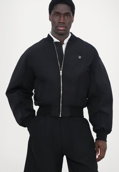Blouson noir en tissu lisse avec fermeture éclair à l'avant, poignets et ourlet côtelés, arborant un petit emblème sur la poitrine.
