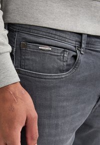Jeans de mezclilla gris con una textura sutil, que presentan un bolsillo frontal y un acento de logo plateado. La mano muestra la textura de la piel y cicatrices.