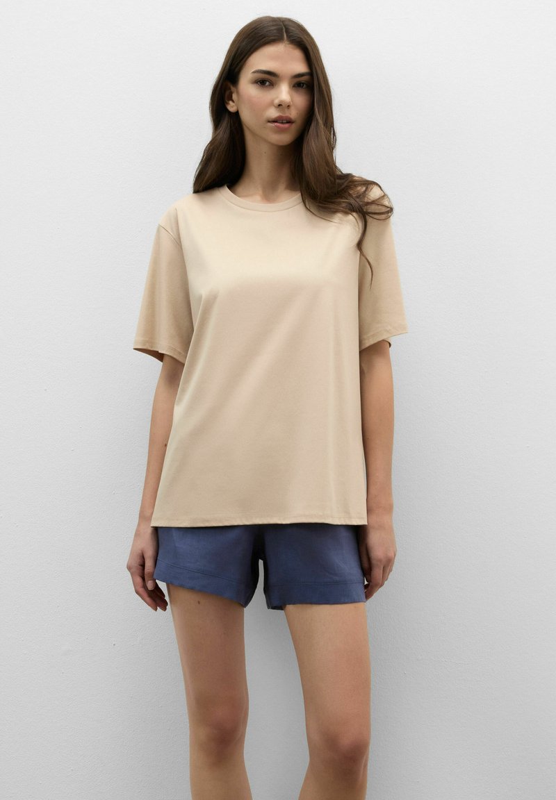 Frau mit langen braunen Haaren, die vor einer weißen Wand steht und ein beiges Kurzarmshirt sowie blaue Shorts trägt.