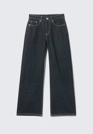 Donkerblauwe jeans met wijde pijpen, knoopsluiting aan de voorkant, rits, lussens voor riem en vijf zakken, tegen een lichte effen achtergrond.