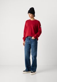 Roter Sweatshirt mit langen Ärmeln, kombiniert mit weit geschnittenen blauen Jeans. Schwarze Mütze und cremefarbene Sneakers mit schwarzen Akzenten. Einfaches Design.