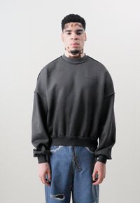 Sweatshirt noir avec un col rond, des épaules tombantes et des poignets côtelés. Associé à un jean bleu déchiré. Le tissu semble doux.