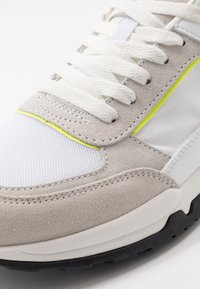 Baskets en daim gris et mesh blanc avec des lacets blancs, présentant un accent jaune néon et une semelle en caoutchouc noire. Tige texturée pour une meilleure adhérence.