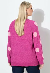 Blonde Frau im Profil, die einen rosa Pullover mit großen hellrosa Punkten auf den Ärmeln trägt, kombiniert mit dunkelblauen Jeans.