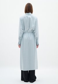 Cămașă lungă din satin de culoare albastră deschisă, cu talie strânsă, mâneci lungi și manșete cu nasturi. Pantaloni negri cu picior larg vizibili în partea de jos.