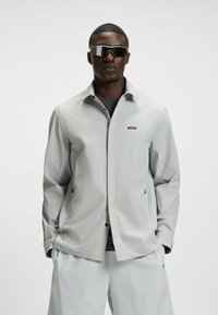 Light gray jacket with logo, featuring a classic collar, button closure, and side pockets. Model wearing dark sunglasses and a gray shirt underneath.