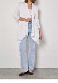 Žena oblečená v bílém ribbeném cardigan s krajkou, bílém topu, světle modrých džínách a tmavě červených balerínách, stojí před jednolitou zdí.