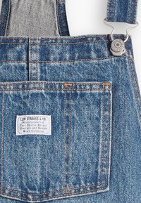 Poche de salopette en denim avec étiquette Levi Strauss & Co., fermoir métallique et double surpiqûre visible sur tissu bleu.