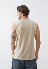 Beige sleeveless cotton shirt with a round neckline, smooth texture, and a tattoo visible on the left arm, paired with blue jeans.