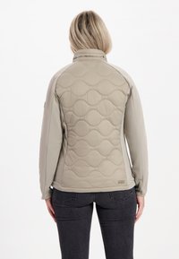 Lätt, beige quiltad jacka med släta ärmar i tyg, upphöjt vågmönster på baksidan och en hög krage för extra värme.