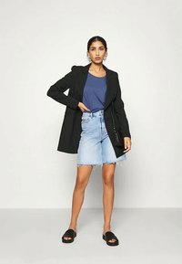 Nainen, jolla on päällään musta pinstripe-takki, sininen t-paita, vaaleat farkkushortsit polven ylle, mustat sandaalit sekä pieni musta laukku.