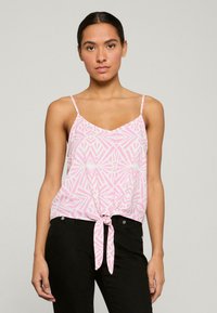 Top in satin rosa con motivi geometrici bianchi, spalline sottili e orlo ancorato davanti, abbinato a pantaloni neri aderenti.