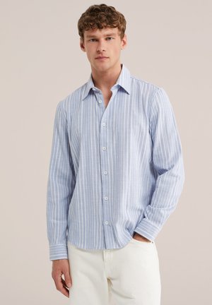 Ung mand iført lyseblå skjorte med lodrette striber og hvide bukser, stående med den ene hånd i lommen mod en ensfarvet baggrund.
