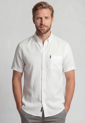 Man met licht haar en baard, gekleed in een wit shirt met korte mouwen en een borstzak, staat met handen in de zakken.