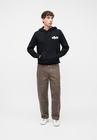 Młody mężczyzna z rękami w kieszeniach, ubrany w czarną bluzę z kapturem, brązowe spodnie i białe trampki, stojący na białym tle.