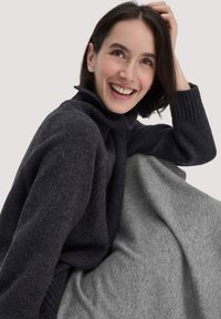 Dunkelgrauer gestrickter Pullover mit hohem Kragen und gerippten Bündchen, geschichtet über einer hellgrauen Decke. Weiche Textur und lässige Passform.