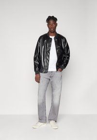 Chaqueta bomber negra con patrón texturizado, camiseta de manga larga gris, jeans grises deslavados y zapatillas blancas con tres franjas laterales.