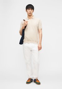 Young man in beige polo, white pants, colorful shoes, carrying navy tote bag over shoulder, standing against plain white background.
