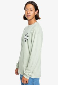 Light green long-sleeve t-shirt in soft material. Features black graphic design on the chest and ribbed cuffs. Worn with blue jeans.