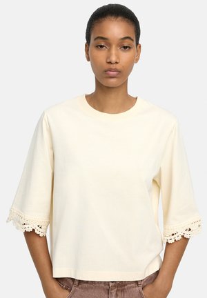 Młoda kobieta o ciemnych włosach związanych do tyłu, ubrana w kremową bluzkę z krótkim rękawem ozdobioną koronkową lamówką na rękawach, z rękami w kieszeniach brązowych spodni.