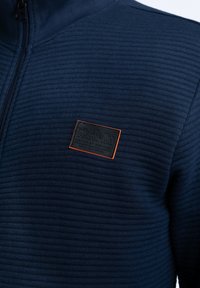 Navy blue zip-up jacket with a ribbed texture, featuring a black leather patch with orange accents on the chest, indicating brand and heritage.