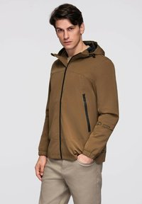 Un jeune homme portant une veste à capuche marron avec des détails de fermeture éclair noirs, se tenant avec une main dans la poche d'un pantalon beige clair, affichant une expression neutre.