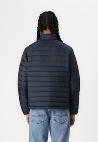 Marineblå dunjakke med lodrette syninger, høj krave og tekstureret stof; iført med lyseblå denimjeans.