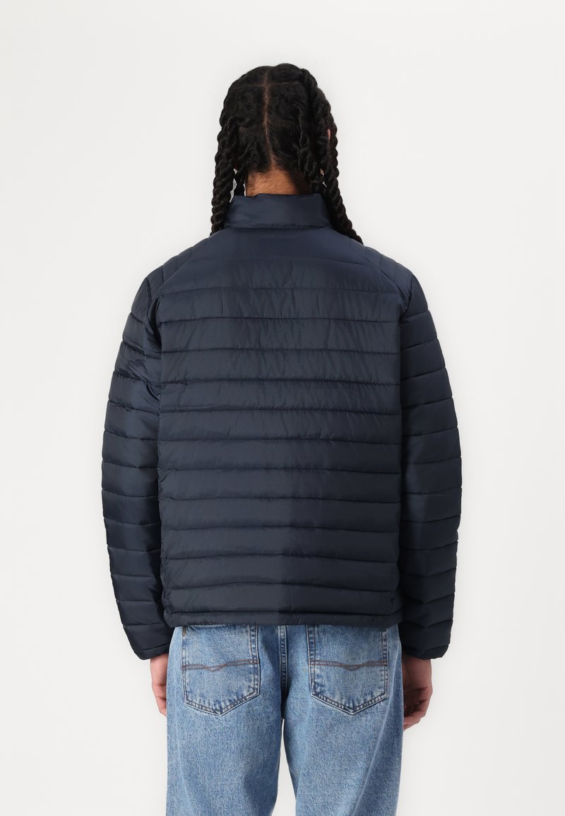 Navyblauwe gewatteerde jas met verticale stiksels, hoge kraag en gestructureerde stof; gedragen met lichtblauwe denim jeans.