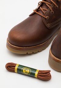 Brune lærstøvler med en teksturert overflate, rund tå og en tan gummi såle, ledsaget av matchende brune lisser pakket inn i grønt emballasje.