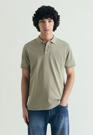 Ung mand med krøllet hår iført lyse olivengrøn polo og blå jeans, stående mod en ensfarvet lys baggrund, med den ene hånd i lommen.