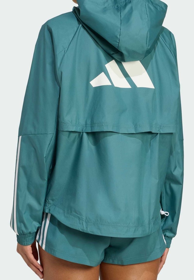Personne portant une veste à capuche bleu sarcelle et un short assorti avec des rayures blanches et un grand logo blanc dans le dos de la veste.