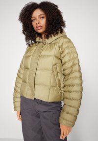 Khaki pufrová bunda s vysokým golierom, prešívanou textúrou a zapínaním na cvoky. Vlastnosti zahŕňajú elastické manžety a skrátený strih.