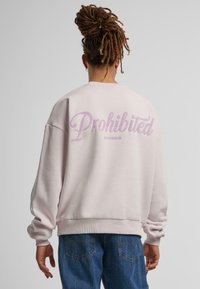 Lys rosa oversized hettegenser med rund hals. "Forbudt" i stor kursiv lilla tekst på baksiden, mykt materiale, ribbede ermer.