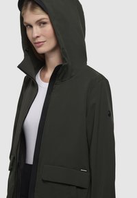 Giacca a cappuccio verde scuro con zip frontale, collo a costine e tasche laterali. Tessuto liscio con finitura opaca e dettaglio con logo sulla manica.