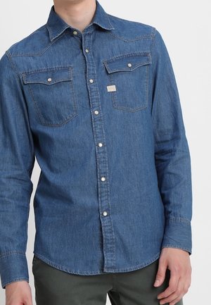 Mann trägt ein langärmeliges blaues Denimhemd mit zwei Brusttaschen und Druckknöpfen.