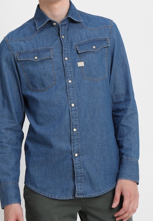 Homme portant une chemise en denim bleu à manches longues, avec deux poches poitrine et des boutons pression.