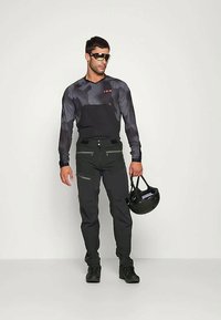 Mężczyzna w czarno-szarym długim rękawie koszulki rowerowej, czarnych spodniach, w okularach przeciwsłonecznych, trzymający czarny kask rowerowy na jednolitym tle.