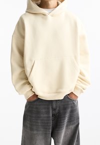 Krémová mikina s klokaní kapsou, měkká textura, volný střih a žebrované manžety, kombinovaná s tmavě šedými, vybledlými džíny.